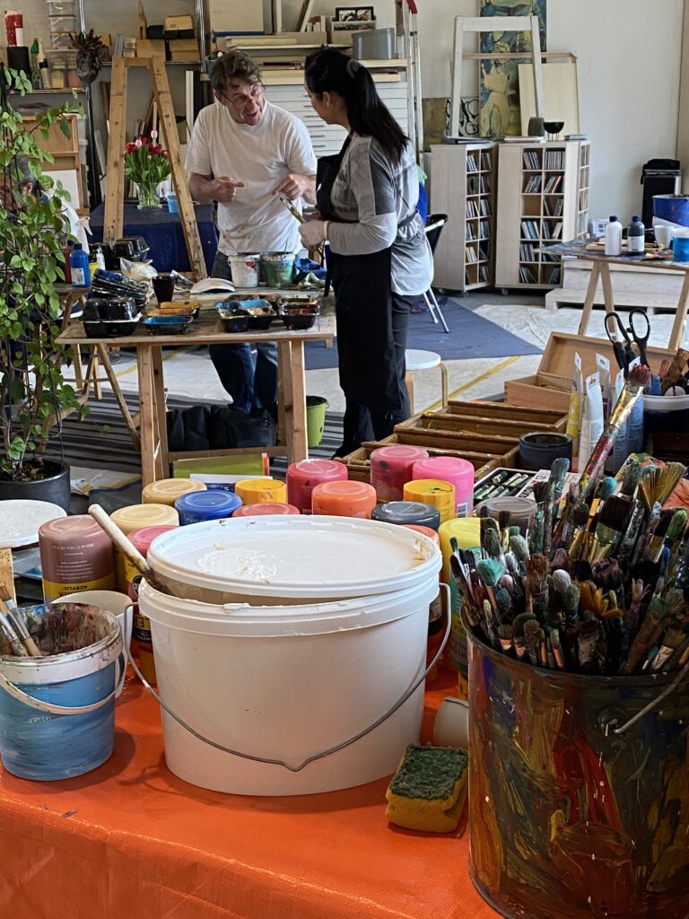 Zwei Personen stehen in einem Kunstatelier an einem Arbeitstisch und sprechen miteinander, umgeben von bunten Farbtöpfen, Pinseln und Malutensilien.