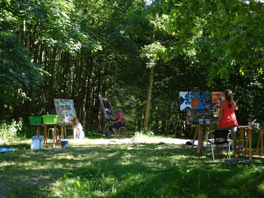 Mehrere Personen malen im Freien an Staffeleien in einer lichtdurchfluteten Waldlichtung; Kunstmaterialien stehen auf Tischen und der Boden ist von Grün umgeben.