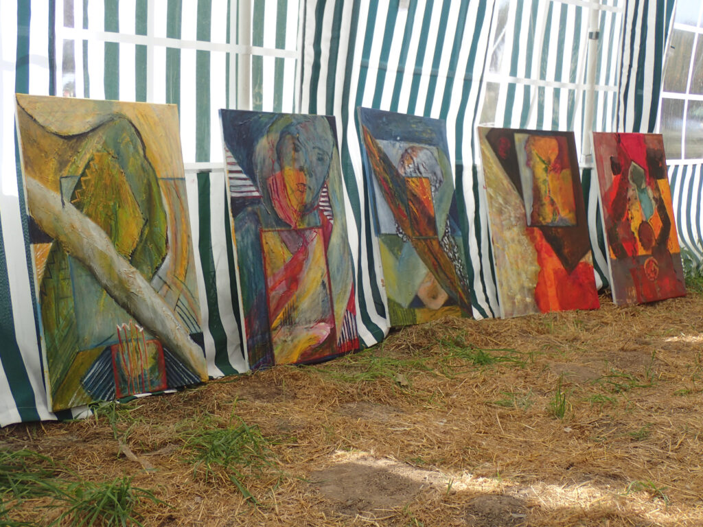 Mehrere abstrakte Gemälde lehnen im Freien an einer gestreiften Zeltwand; die farbintensiven Arbeiten zeigen unterschiedliche Formen, Strukturen und Kompositionen.