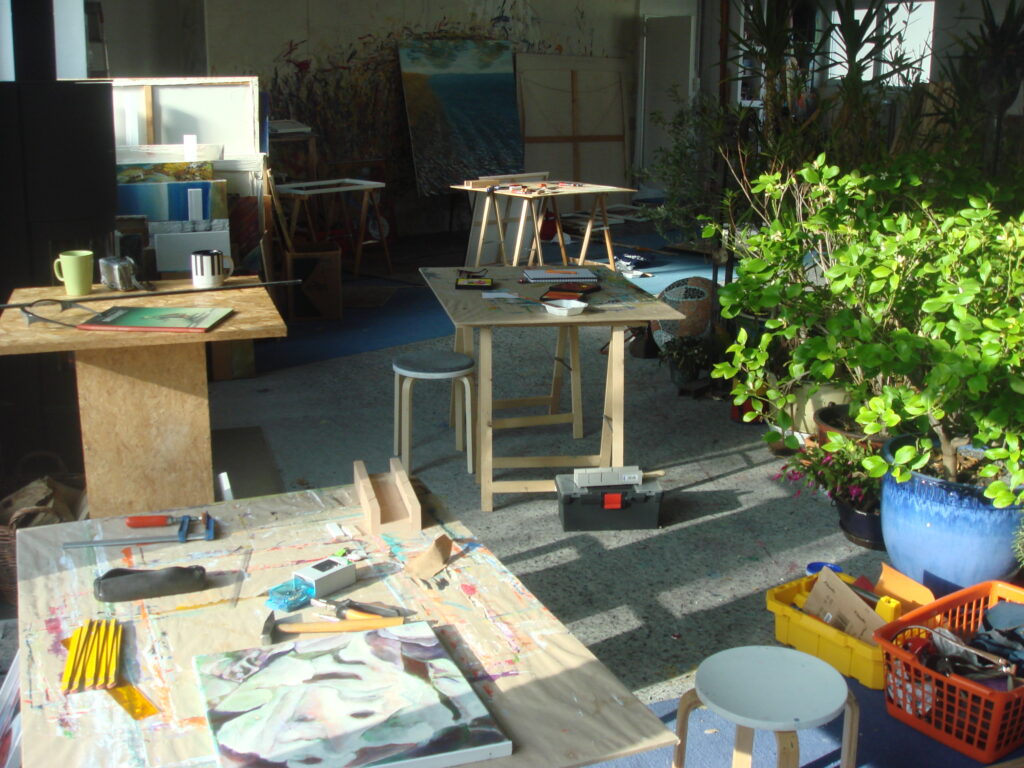 Heller Arbeitsbereich im Atelier mit Tischen voller Malutensilien, Werkzeugen und Skizzen; im Hintergrund stehen Staffeleien und Pflanzen, die eine inspirierende und lebendige Atmosphäre schaffen.