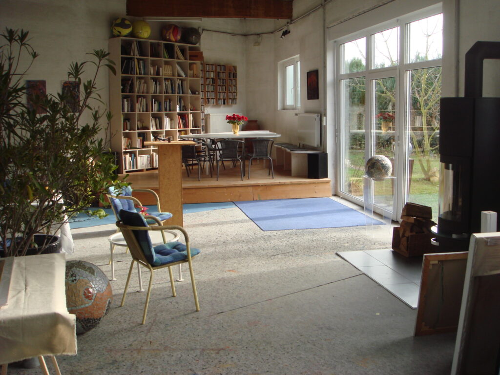 Lichtdurchfluteter Atelierraum mit Sitzbereich, Bücherregalen und einem erhöhten Tischplatz; große Glasfronten öffnen den Blick in den Garten und schaffen eine ruhige, einladende Atmosphäre.