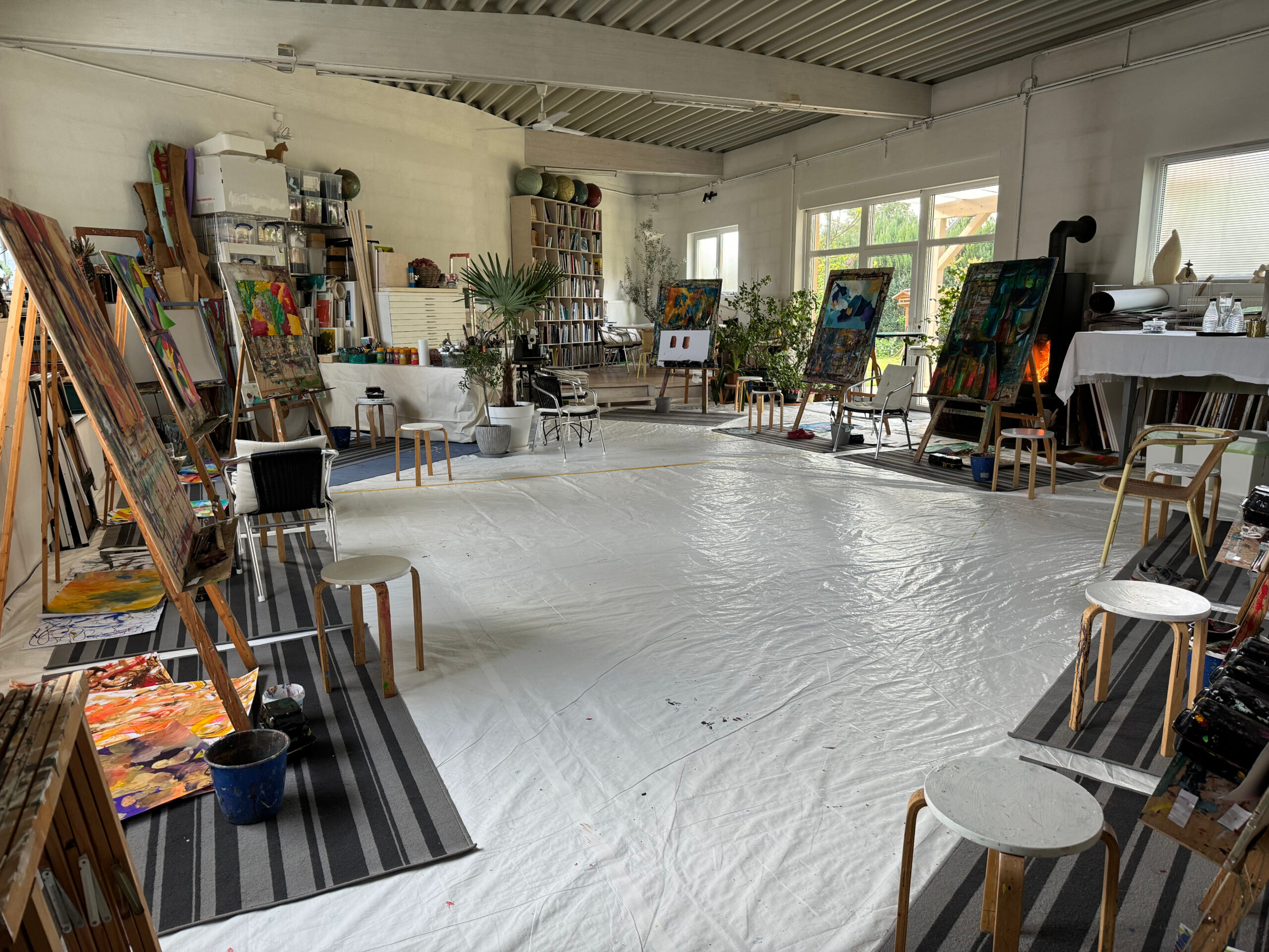 Großer, lichtdurchfluteter Atelierraum mit Staffeleien, farbenreichen Bildern und Malmaterialien; die offene Gestaltung und Pflanzen schaffen eine inspirierende Arbeitsatmosphäre für kreative Kurse.