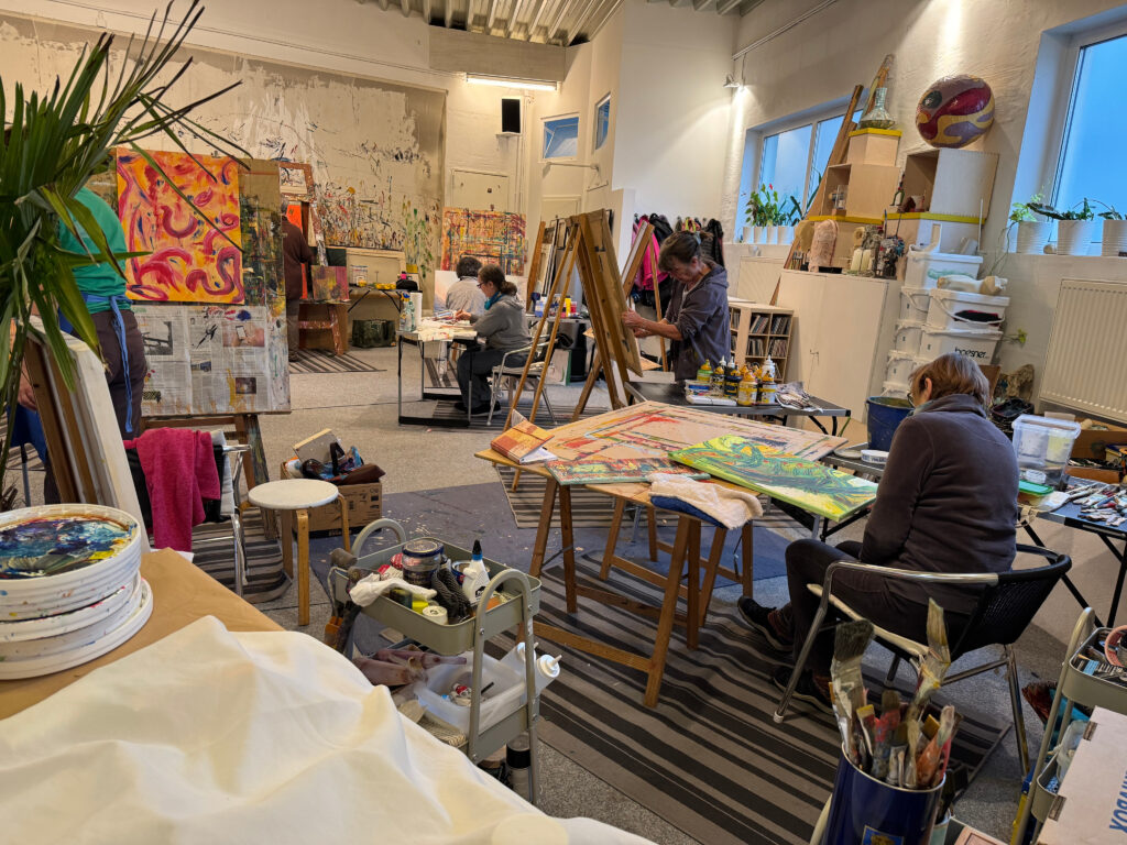 Mehrere Personen arbeiten konzentriert an Staffeleien und Tischen in einem großzügigen Atelierraum; farbenfrohe Leinwände, Malutensilien und eine kreative Arbeitsatmosphäre füllen den Raum.