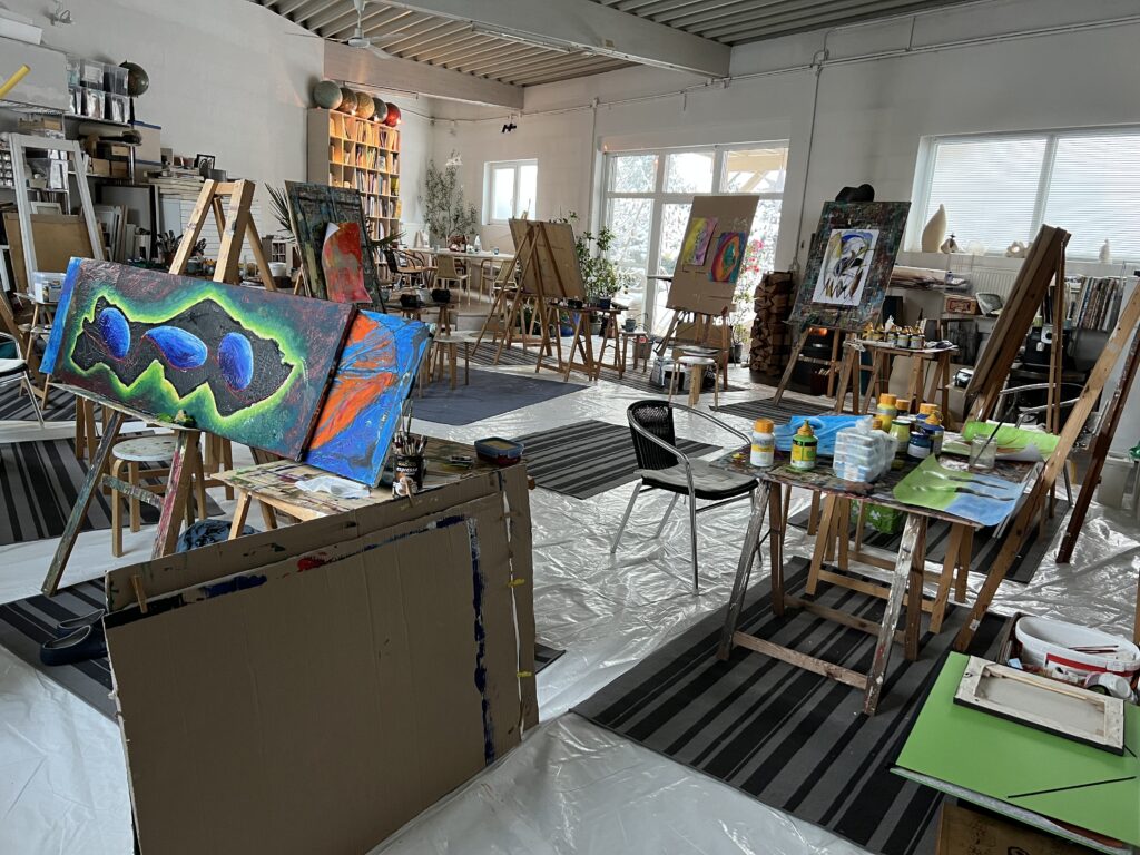 Großer, gut beleuchteter Atelierraum mit mehreren Staffeleien, farbintensiven Leinwänden und Tischen voller Malmaterialien; die offene und kreative Umgebung lädt zum Arbeiten und Experimentieren ein.
