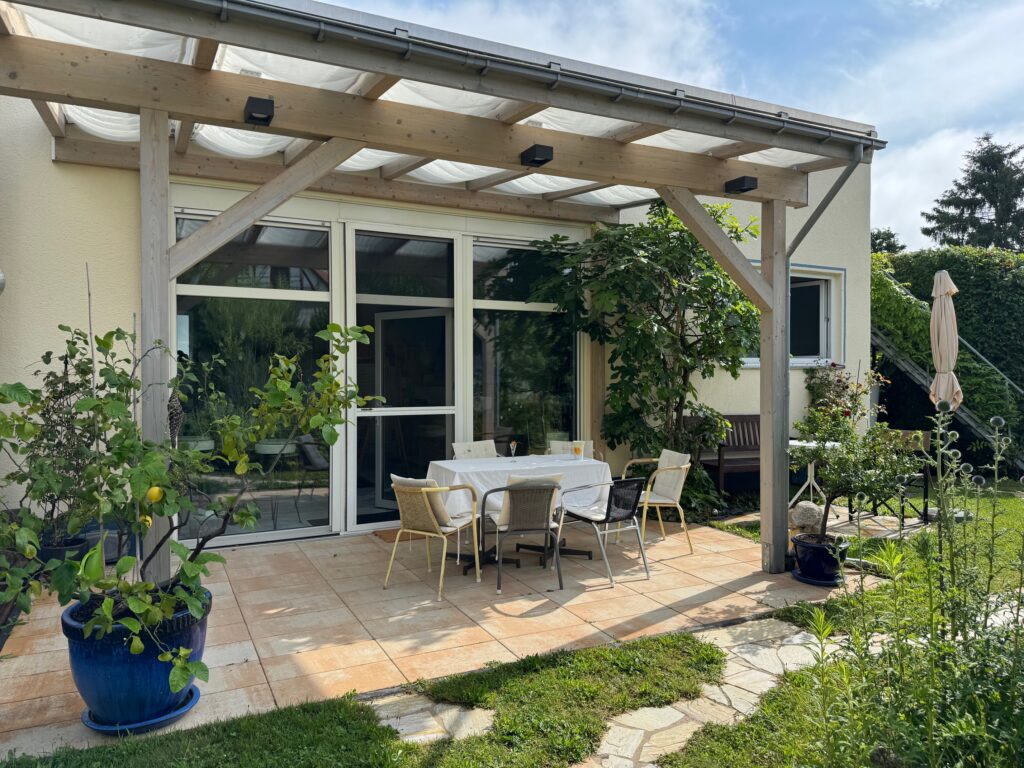 Gemütliche Terrasse vor dem Atelier mit überdachtem Sitzbereich, Pflanzen und einladender Atmosphäre; der Eingang führt in den lichtdurchfluteten Atelierraum.