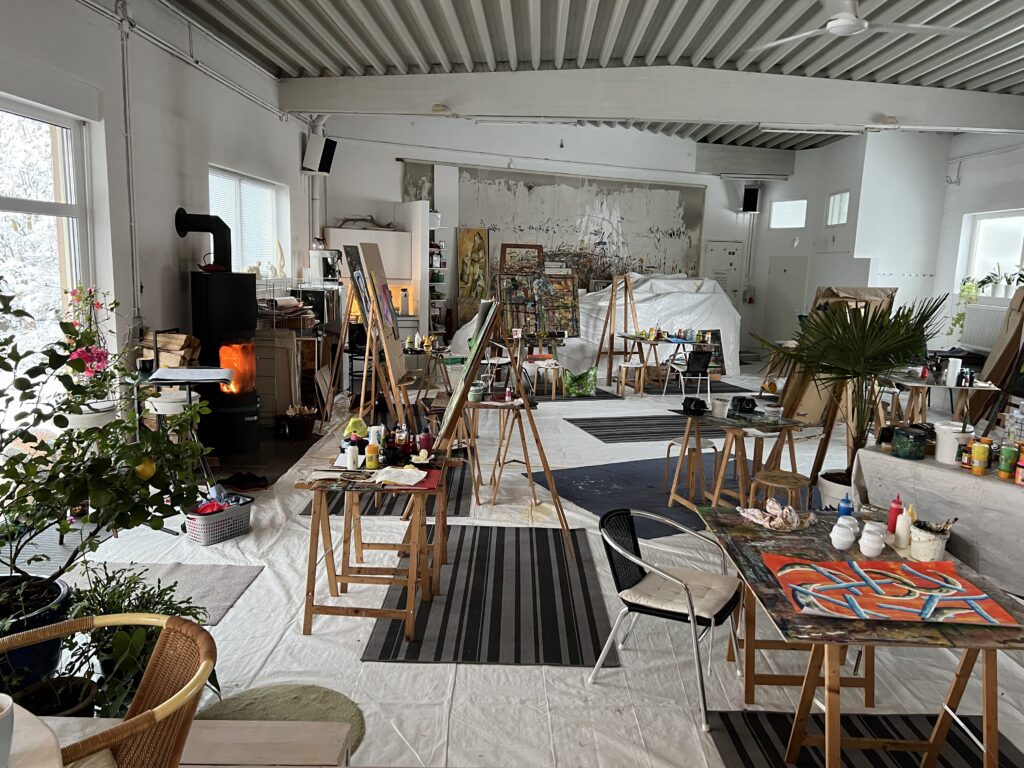 Weitläufiger Atelierraum mit zahlreichen Staffeleien, Malmaterialien und Pflanzen; durch die Fenster fällt winterliches Licht, während im Hintergrund ein Kaminofen ein warmes, einladendes Ambiente schafft.