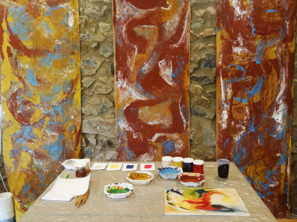 Große bemalte Papierbahnen in warmen Rot-, Gelb- und Blautönen hängen vor einer Steinwand. Davor steht ein Arbeitstisch mit Farbschalen, Pinseln und kleinen Farbkarten – eine vorbereitete Malstation im Atelier Freifarbe.