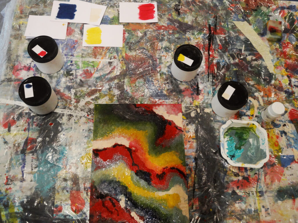 Auf einer bunt befleckten Arbeitsunterlage liegen mehrere Farbkarten, Farbgläser und eine abstrakte Farbstudie; die Szene zeigt verschiedene Pigmenttests und das Experimentieren mit intensiven Farbkombinationen.