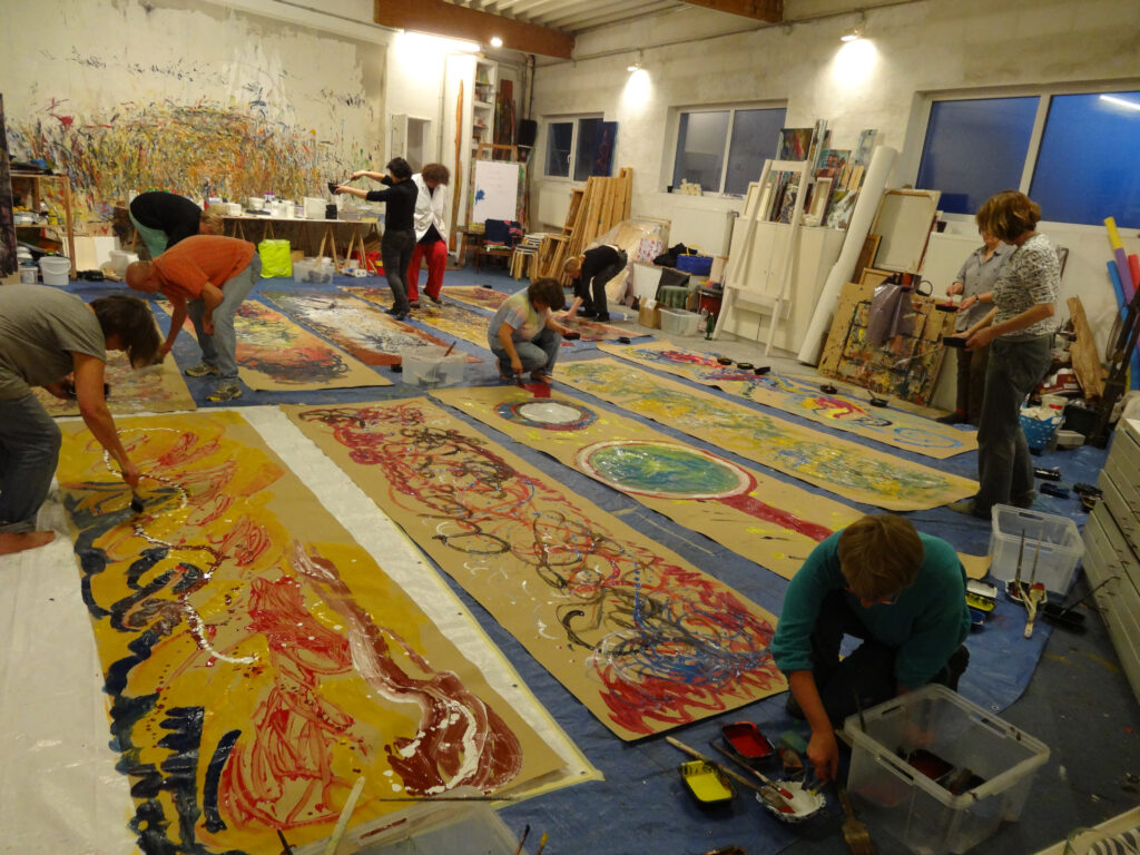 Mehrere Teilnehmende arbeiten gemeinsam an großformatigen Papierbahnen, die am Boden des Ateliers ausgelegt sind; kräftige Farben, dynamische Gesten und eine lebendige Gruppenatmosphäre prägen den kreativen Prozess.