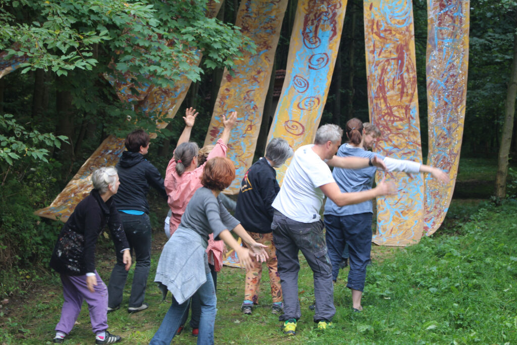 Teilnehmende eines Kunstworkshops bewegen sich gemeinsam vor mehreren großformatigen, bemalten Papierbahnen, die zwischen Bäumen hängen; die Gruppe wirkt dynamisch und eingebunden in eine kreative Aktion im Wald.