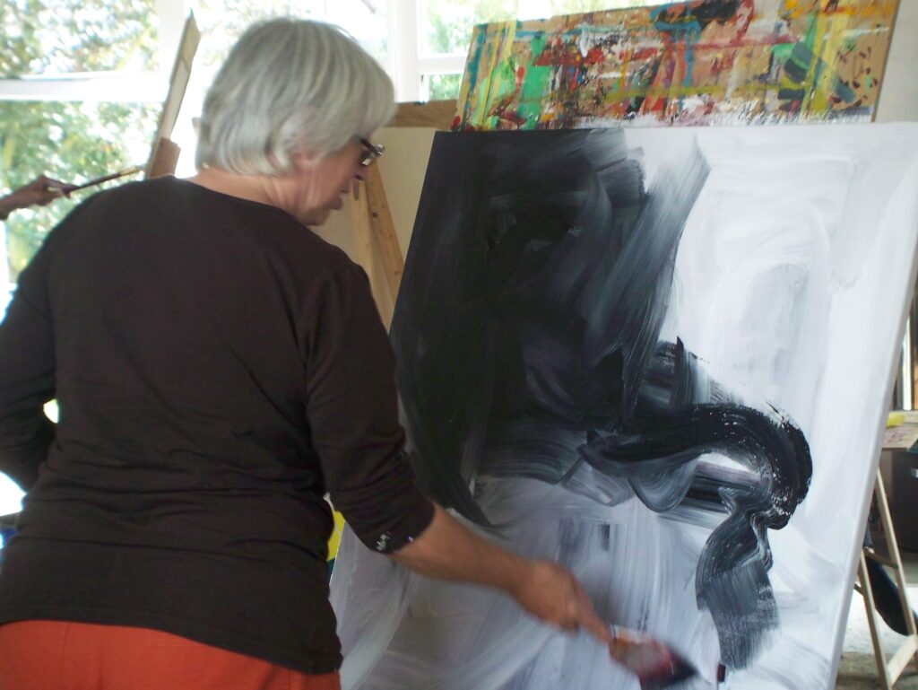 Eine Person arbeitet an einer großformatigen Leinwand mit dynamischen Schwarz-Weiß-Pinselstrichen; die Szene zeigt einen intensiven kreativen Moment im Atelier.