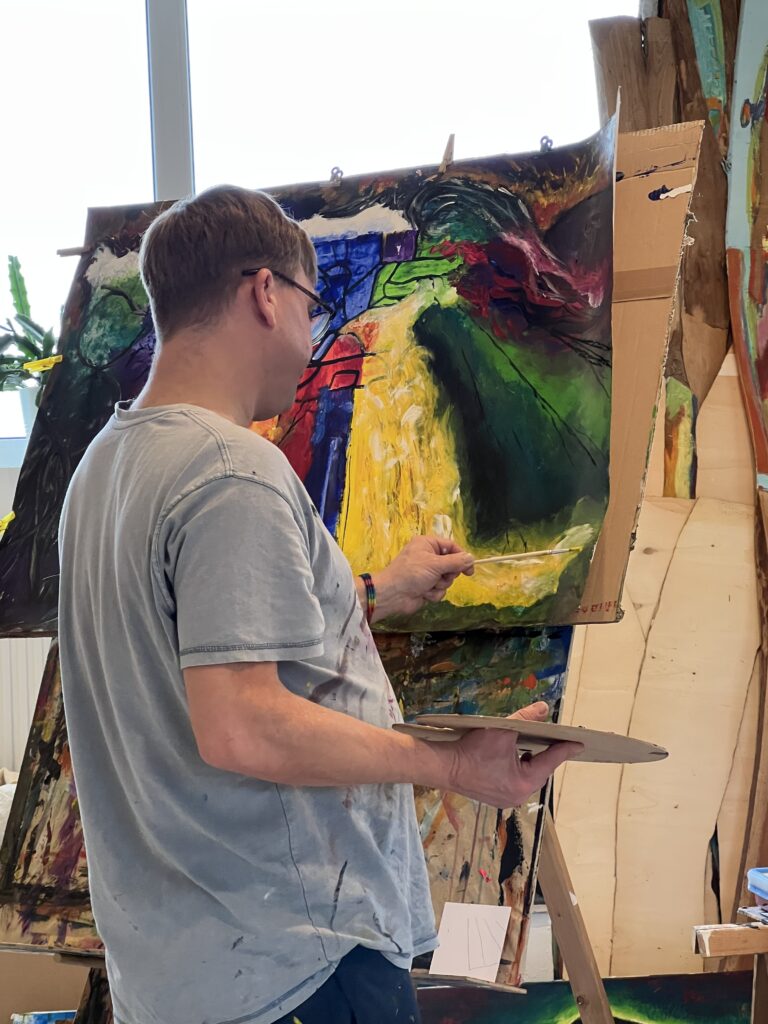 Eine Person malt konzentriert an einer farbintensiven abstrakten Leinwand; kräftige Gelb-, Grün-, Blau- und Rottöne sowie die Atelierumgebung unterstreichen die kreative Arbeitssituation.