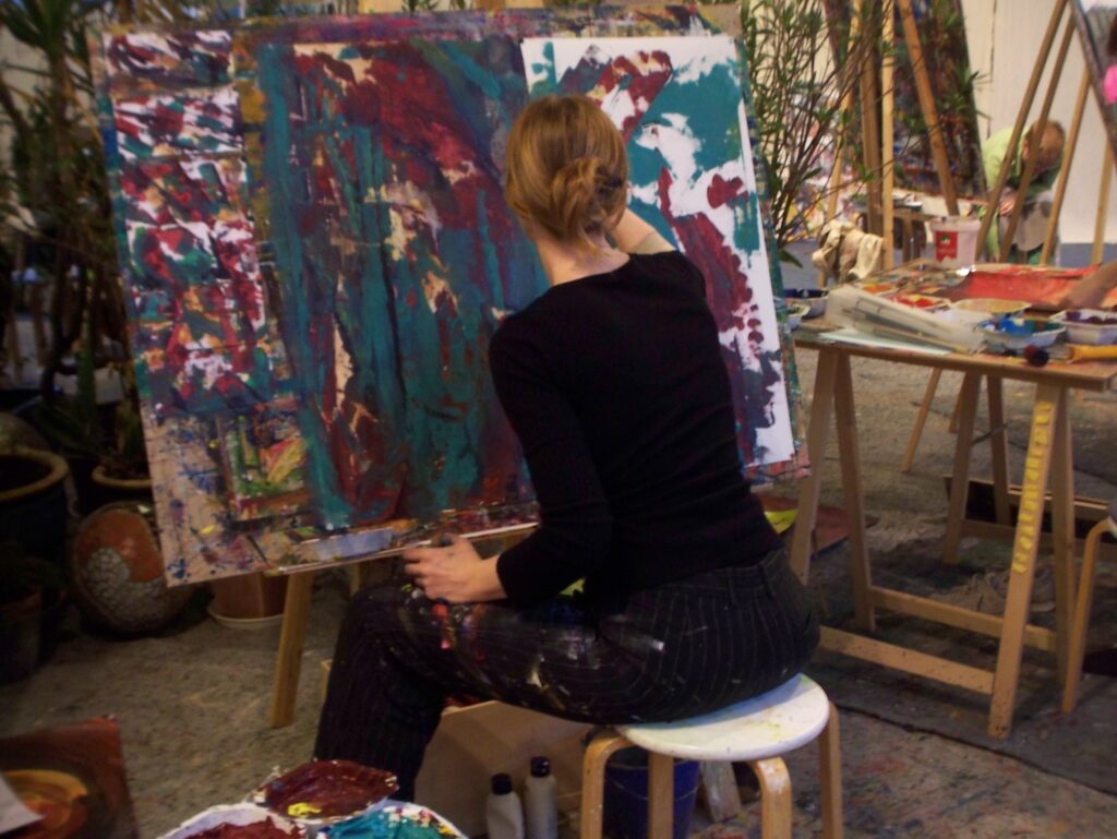 Eine Person sitzt auf einem Hocker und arbeitet konzentriert an einer großformatigen abstrakten Leinwand; farbintensive Pinselstriche und zahlreiche Malmaterialien umgeben die kreative Szene im Atelier.
