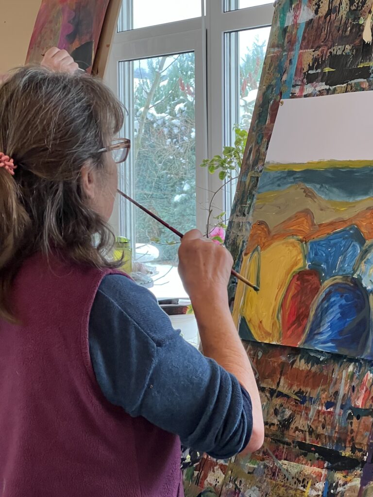 Eine Person malt an einer Staffelei eine farbintensive abstrakte Landschaft; durch die großen Fenster fällt Winterlicht ins Atelier und schafft eine ruhige, konzentrierte Stimmung.