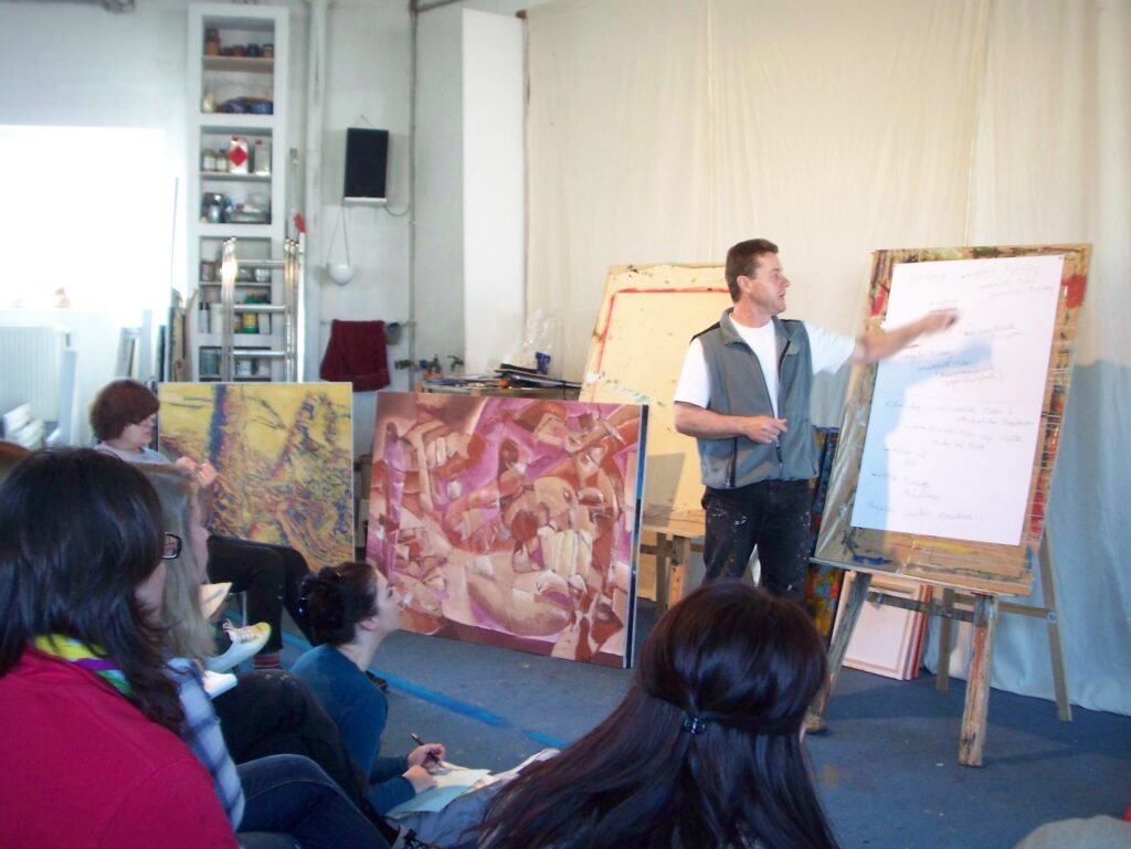 Ein Dozent erklärt vor einer Gruppe von Teilnehmenden grundlegende Aspekte des Bildaufbaus an einem Flipchart; im Hintergrund stehen großformatige Gemälde, die den kreativen Unterricht im Atelier ergänzen.