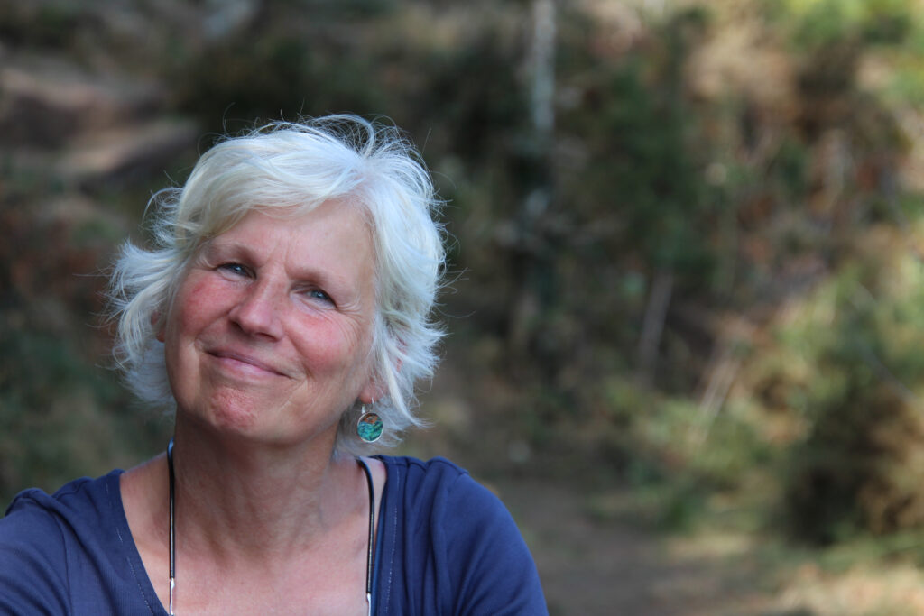 Porträt einer lächelnden Frau (Heidi Schrickel) mit weißem Haar in natürlicher Umgebung; sie wirkt freundlich und zugewandt und sitzt entspannt im Freien.